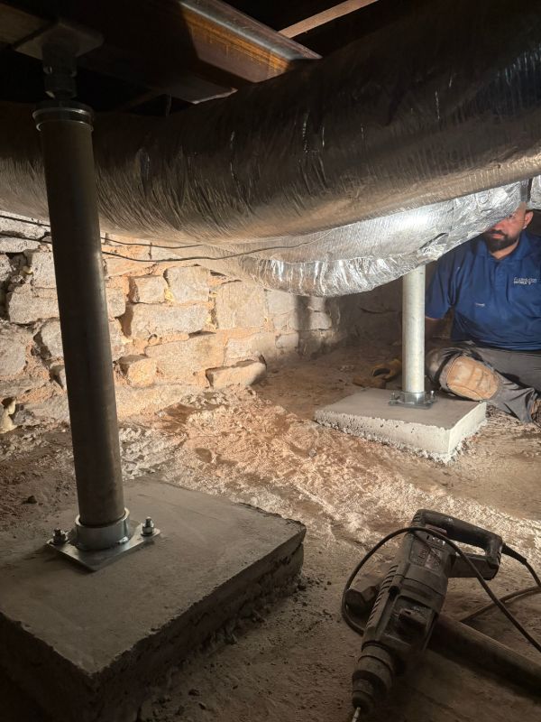Steel support posts set on new concrete footing pads in a crawl space beside a stone foundation wall