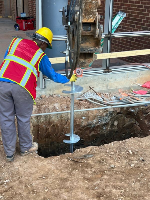 Crew member in safety gear guiding a helical pile installation beside a commercial building